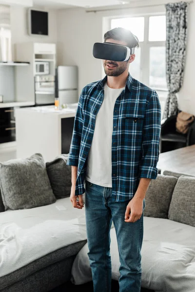 Homem de camisa quadriculada usando fone de ouvido de realidade virtual perto do sofá em casa — Fotografia de Stock