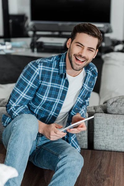 Selective focus of handsome man smiling at camera while holding digital tablet on floor in living room — Stock Photo
