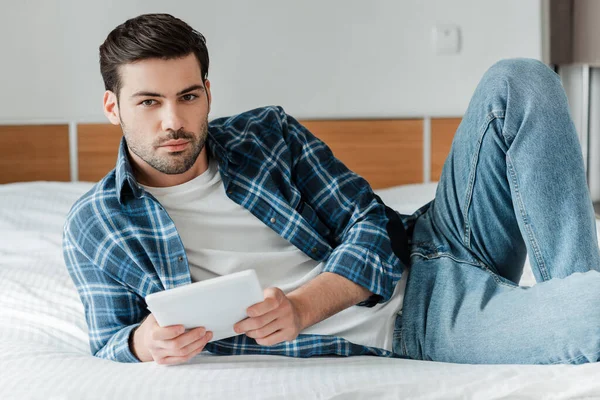 Handsome man in plaited shirt and jeans holding digital tablet ans looking at camera on bed — Stock Photo