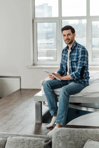 Selektiver Fokus eines gutaussehenden Mannes im karierten Hemd, der in die Kamera blickt, während er ein digitales Tablet auf dem Bett hält — Stockfoto