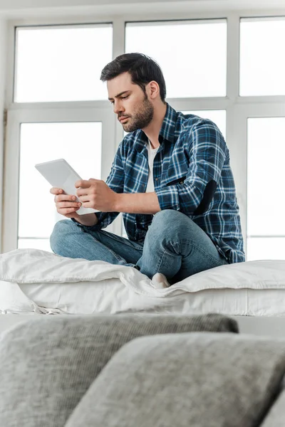 Enfoque selectivo del hombre usando tableta digital mientras está sentado en la cama cerca del sofá en casa - foto de stock