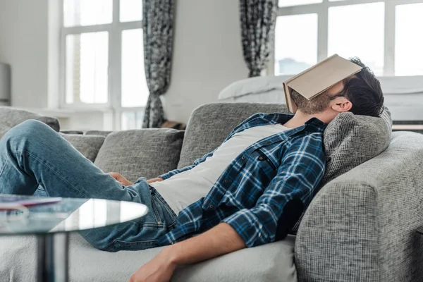 Concentration sélective de l'homme couché sur le canapé avec livre sur le visage à la maison — Photo de stock