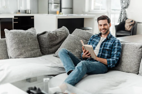 Selective focus of handsome man smiling at camera while reading book on couch at home — Stock Photo