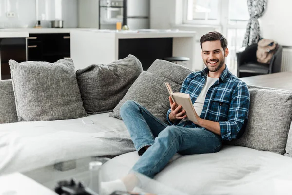 Enfoque selectivo del hombre guapo sonriendo a la cámara mientras lee el libro en el sofá en casa - foto de stock