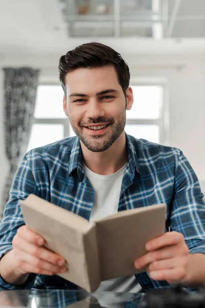 Foco seletivo de homem bonito na camisa entranhada sorrindo para a câmera enquanto segura o livro em casa — Fotografia de Stock