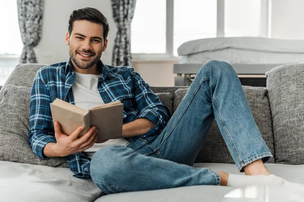 Selektiver Fokus eines Mannes, der in die Kamera lächelt, während er sein Buch zu Hause auf dem Sofa hält — Stockfoto
