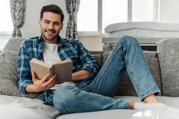 Foco seletivo do homem sorrindo para a câmera enquanto segura o livro no sofá em casa — Fotografia de Stock