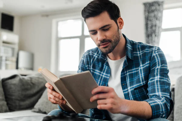 Enfoque selectivo del hombre guapo en camisa trenzada libro de lectura en casa - foto de stock