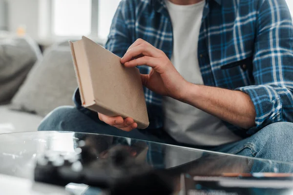 Cropped view of man in plaited shirt and jeans holding book on couch at home — Stock Photo