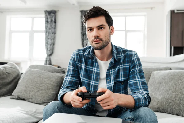 KYIV, UCRANIA - 13 DE ABRIL DE 2020: Hombre guapo con camisa trenzada jugando videojuego cerca de la computadora portátil en la mesa de café - foto de stock