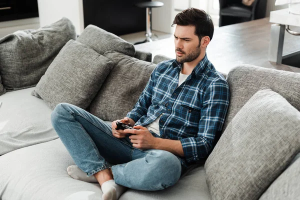 KYIV, UKRAINE - APRIL 13, 2020: Handsome man holding joystick while playing video game on sofa at home — Stock Photo