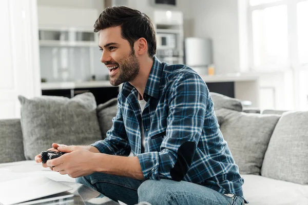 KYIV, UCRANIA - 13 DE ABRIL DE 2020: Hombre sonriente sosteniendo el joystick mientras juega un videojuego cerca de la computadora portátil en la mesa de café - foto de stock