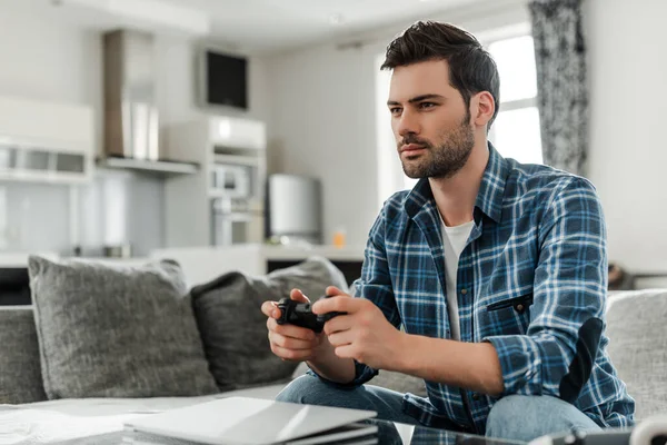 KIEW, UKRAINE - 13. April 2020: Selektiver Fokus eines gutaussehenden Mannes mit Joystick, während er zu Hause auf der Couch sitzt — Stockfoto