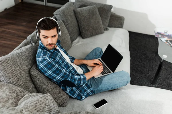 Высокоугольный вид человека в наушниках с ноутбуком рядом со смартфоном на диване — стоковое фото
