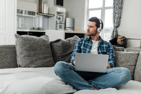 Bello freelance in cuffia con computer portatile sul divano di casa — Foto stock