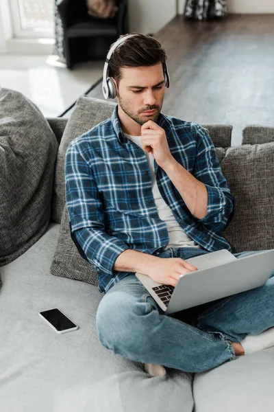 Handsome freelancer in headphones using laptop near smartphone with blank screen on couch — Stock Photo