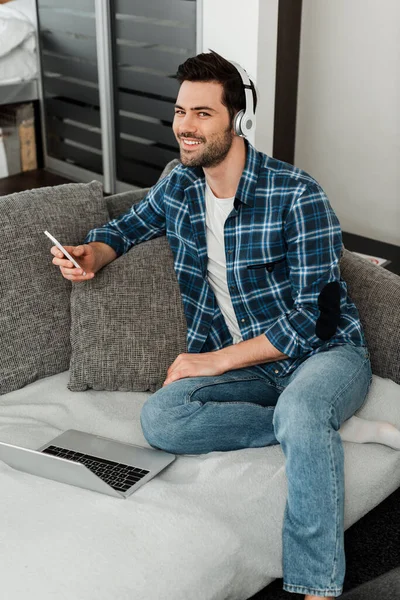 Sorrindo homem em fones de ouvido segurando smartphone perto de laptop no sofá em casa — Fotografia de Stock