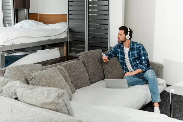 Selektiver Fokus eines gutaussehenden Mannes mit Kopfhörern, der sein Smartphone in der Nähe des Laptops auf der heimischen Couch hält — Stockfoto