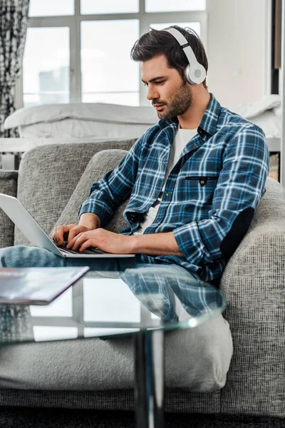 Selective focus of freelancer in plaited shirt using headphones and laptop on couch — Stock Photo