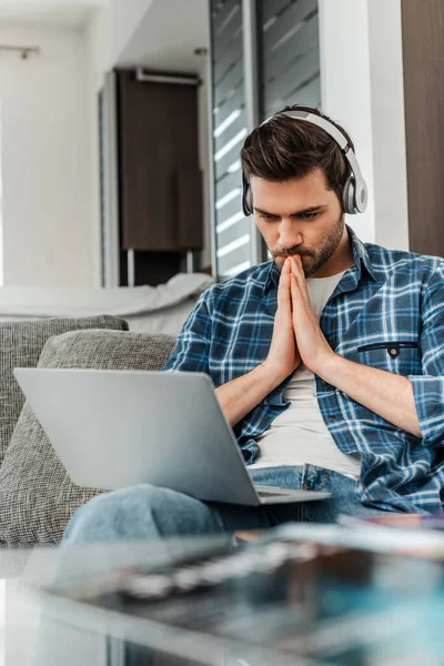 Selective focus of freelancer in headphones with prayer hands looking at laptop on couch — Stock Photo