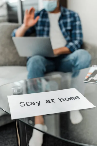 Selective focus of card with stay at home lettering and man in medical mask having video call on laptop at home — Stock Photo