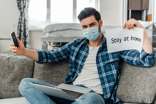 Мужчина в медицинской маске держит смартфон, карточку с домашними надписями и ноутбук на диване — стоковое фото