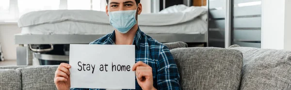 Orientação panorâmica do homem em cartão de retenção máscara médica com estadia em casa lettering no sofá em casa — Fotografia de Stock