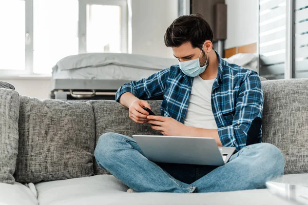 Freelancer em máscara médica usando smartphone e laptop no sofá em casa — Fotografia de Stock