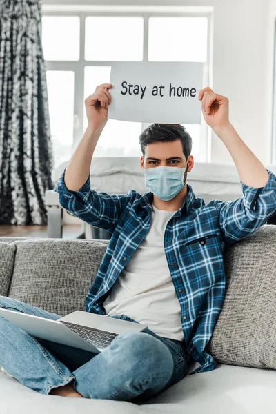 Freelance en masque médical tenant la carte avec rester à la maison lettrage près de l'ordinateur portable sur le canapé — Photo de stock