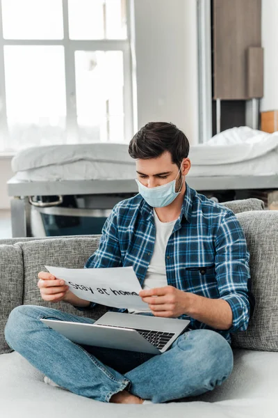 Freelancer en tarjeta de retención de máscara médica con letras de estancia en casa y computadora portátil en el sofá - foto de stock