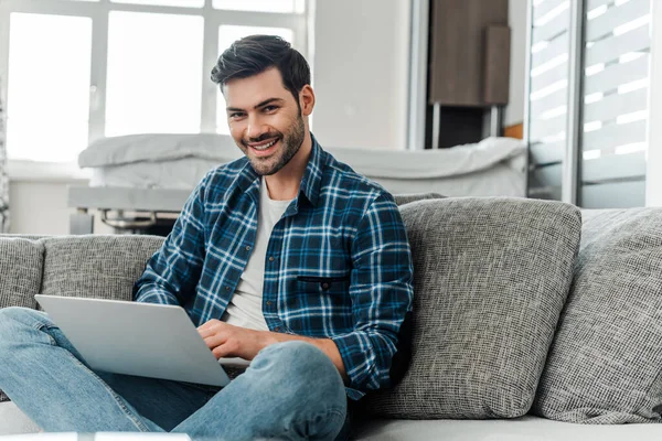 Handsome freelancer smiling at camera while using laptop at home — Stock Photo
