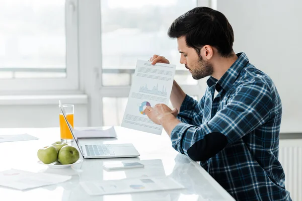 Вид сбоку человека, указывающего на бумагу с графиками возле ноутбука и аппликациями на столе на кухне — стоковое фото