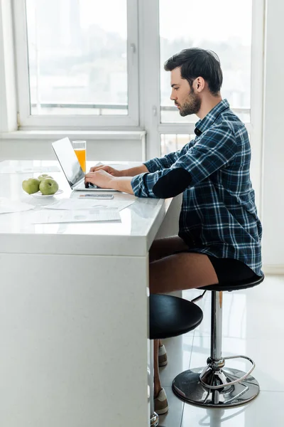 Vue latérale du pigiste en chemise et culotte à l'aide d'un ordinateur portable près des documents sur la table dans la cuisine — Photo de stock