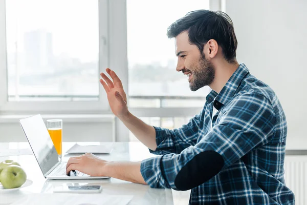 Vista lateral do freelancer sorrindo ter chamada de vídeo no laptop perto de maçãs e vidro de suco de laranja na mesa da cozinha — Fotografia de Stock