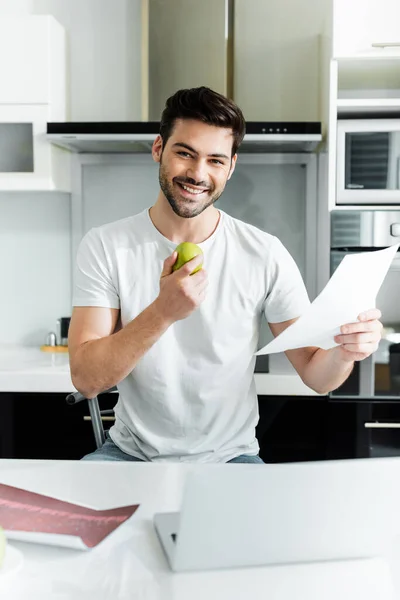 Selective focus of smiling freelancer holding apple and document near laptop on table in kitchen — Stock Photo