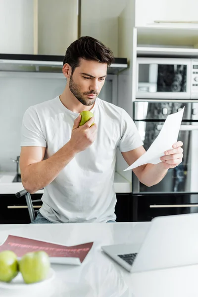 Foco seletivo de homem bonito trabalhando com papéis e segurando maçã perto de laptop na cozinha — Fotografia de Stock