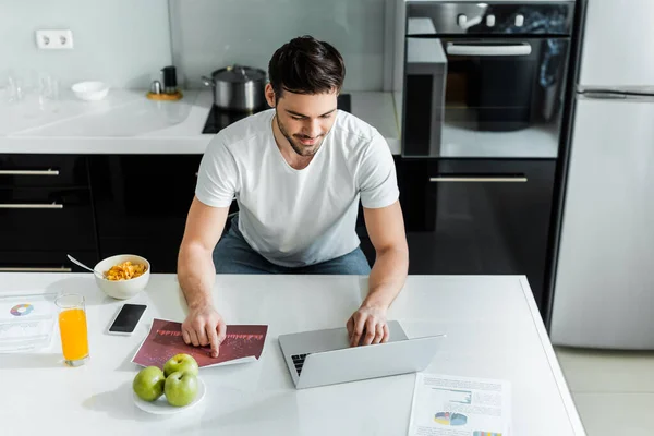 Hombre guapo usando el ordenador portátil mientras trabaja con gráficos en papeles cerca de cereales y manzanas en la mesa de la cocina - foto de stock