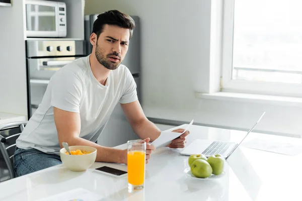 Libero professionista pensieroso che guarda la macchina fotografica mentre tiene la carta vicino a aggeggi e cereali sul tavolo della cucina — Foto stock