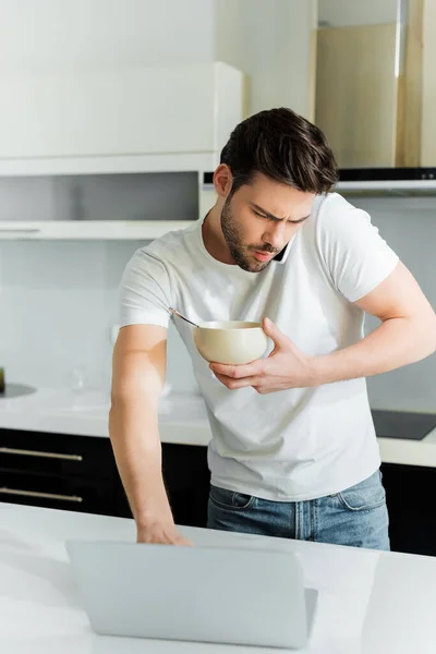 Foco seletivo do homem falando no smartphone, usando laptop e tigela na cozinha — Fotografia de Stock