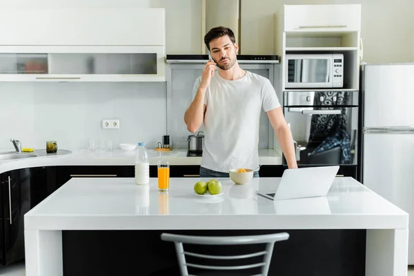 Man talking on smartphone near laptop, cereals and orange juice on kitchen table — Stock Photo