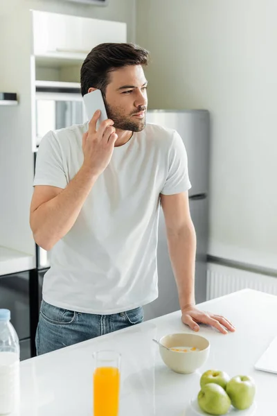 Selektiver Fokus eines gutaussehenden Mannes, der neben Müsli, Äpfeln und Orangensaft auf dem Tisch mit dem Smartphone spricht — Stockfoto