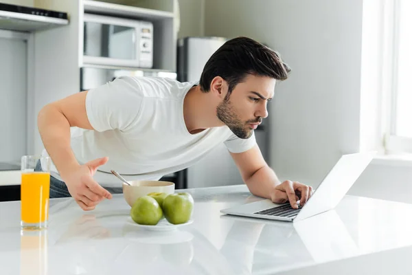 Foco seletivo do homem usando laptop perto de suco de laranja, maçãs e tigela na mesa da cozinha — Fotografia de Stock