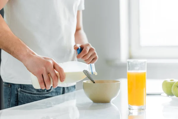 Vista recortada del hombre vertiendo leche en un tazón cerca del jugo de naranja y manzanas en la mesa - foto de stock