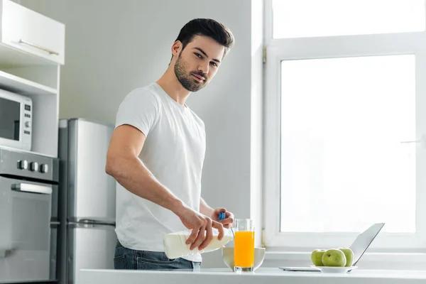 Man looking at camera while pouring milk in bowl near laptop, apples and orange juice on table in kitchen — Stock Photo