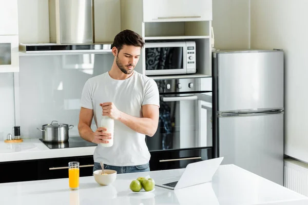 Bell'uomo che tiene una bottiglia di latte vicino al computer portatile e cereali sul tavolo in cucina — Foto stock