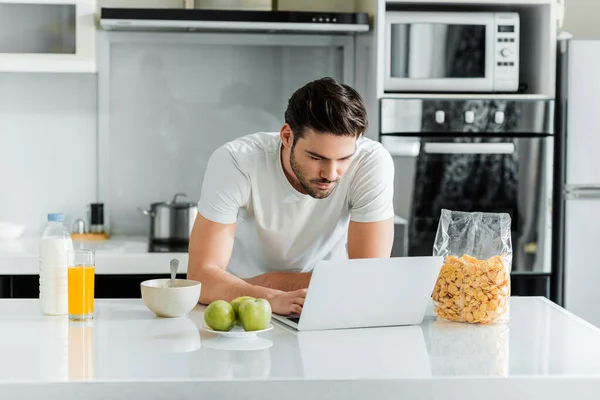 Человек с ноутбуком рядом с зерновыми и апельсиновым соком на кухонном столе — стоковое фото