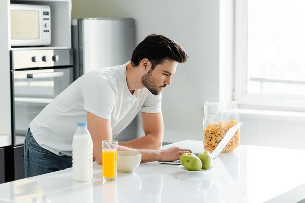 Homme utilisant un ordinateur portable près des céréales, du lait et du jus d'orange sur la table de cuisine — Photo de stock