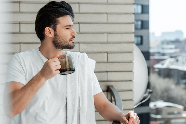 Selective focus of handsome man with towel on shoulder holding cup of coffee on balcony — Stock Photo