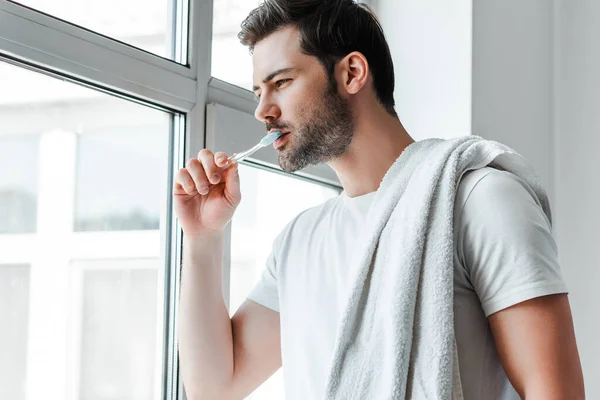 Handsome man with towel on shoulder brushing teeth and looking at window — Stock Photo