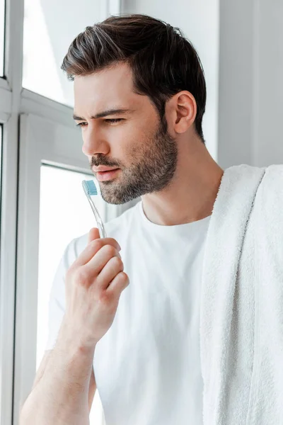 Hombre barbudo sosteniendo toalla y cepillo de dientes mientras mira a la ventana en casa - foto de stock